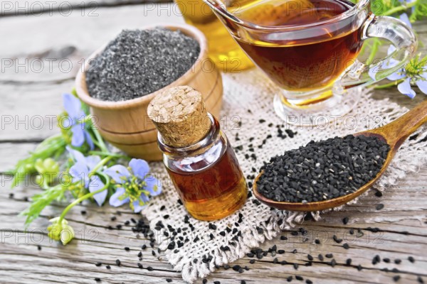 Nigella sativa oil in vial, gravy boat and bottle, seeds in a spoon and black cumin flour in a bowl on burlap, twigs with blue flowers and leaves on background of an old wooden board