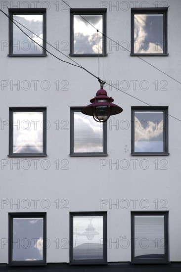 A hanging lamp in front of the façade of a residential building with windows reflecting the clouds
