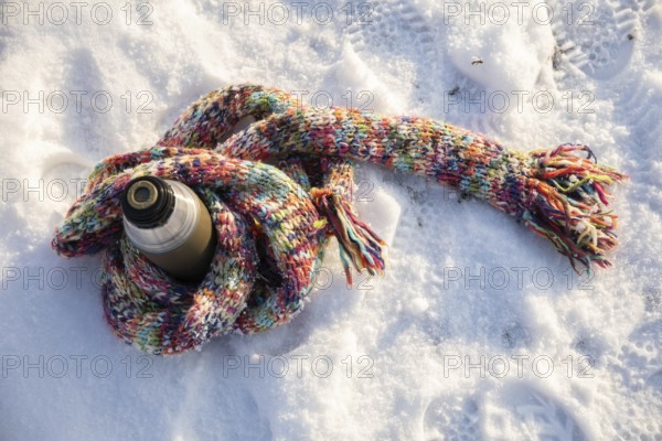 A long, colorful wool scarf lies on the snow. The scarf is wrapped around a thermos. The thermos is standing on the snow. Fresh shoe marks can be seen around the thermos and the scarf