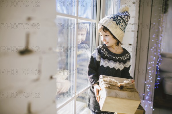 Child sitting at the window on a sunny Christmas day, making presents wrapped in boxes in paper. dressed in knitted warm woollen clothes and hat. Inside the house with a stylish interior