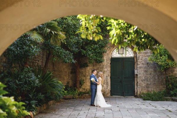 The bride and groom stand hugging in the coutryard in the old town of Perast, view through the arch.