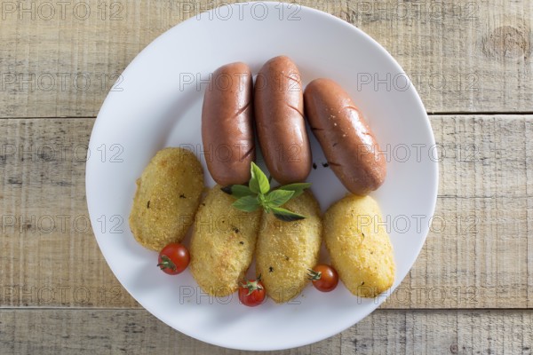 Pork sausages and breaded potatoes fried in a dish and decorated with cherry tomato placed on wooden table