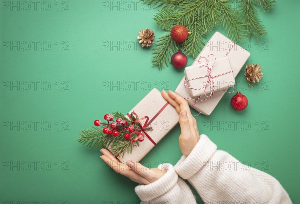 Christmas or New Year celebration green paper festive background with female hands holding wrapped present box, decoration fir tree, cones, berries, sparkly red balls. Space for text, food photography, food photography