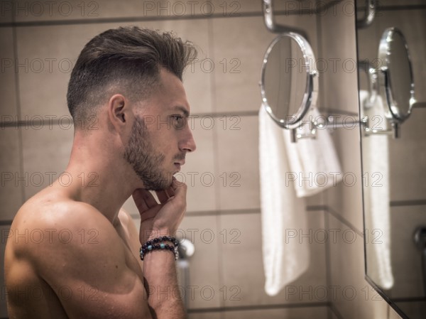 Shirtless muscular handsome young man looking at himself in bathroom mirror in the morning