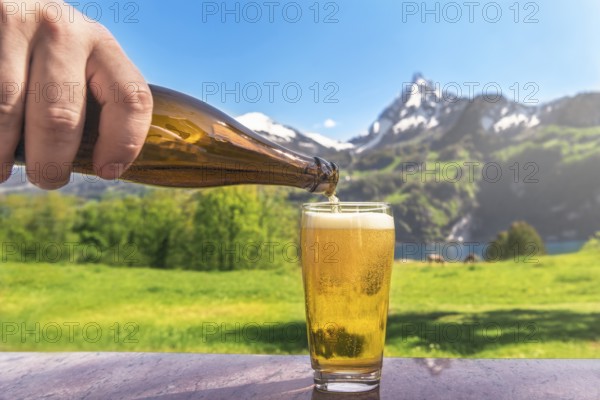 Hand of a man pouring beer from a bottle in a glass with a summer scenery in the background with mountains and green meadows, on a sunny day