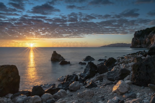 A colorful sunrise at Cala Gonone with black and white rocks and boulders and a sunburst