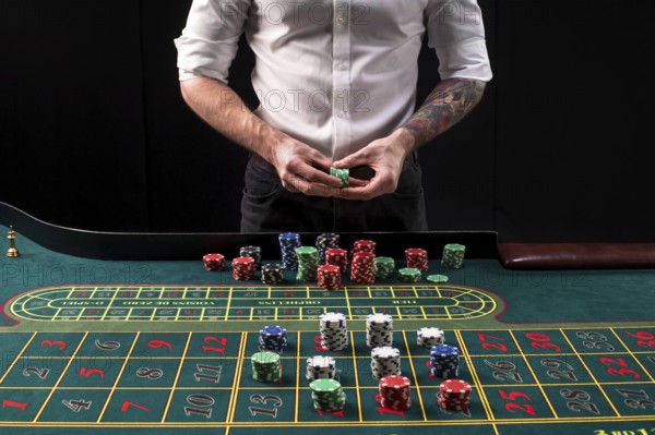 A close-up vibrant image of green casino table with roulette, with the hands of croupier and multicolored chips, on black background. Casino. Gambling. Roulette. Betting