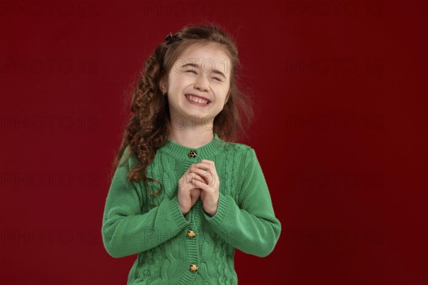 Portrait of a laughing tiny kid with a long, curly hair looking happy. Charming girl in a knitted green sweater is posing against a red background. Sincere emotions concept