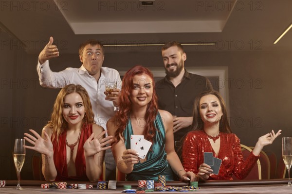 Happy friends are playing poker at casino. They are celebrating their win, smiling and posing at the table against a dark smoke background. Cards, chips, money, alcohol, fortune, gambling, entertainment concept