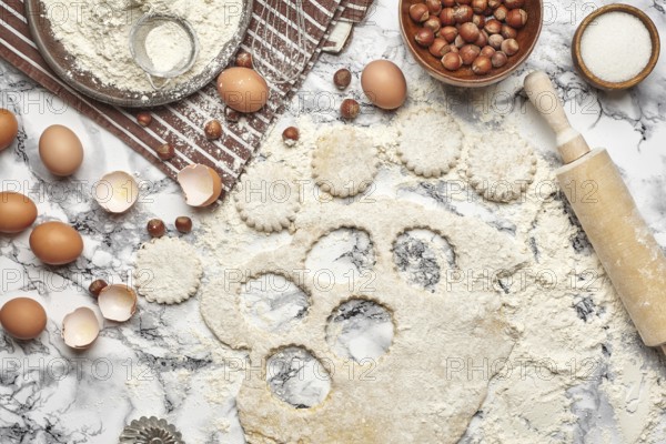 Close-up shot. Top view of a kitchenware and baking ingredients on a marble table background. There are some eggs and an eggshell, flour on a brown apron, sieve, sugar, scattered hazelnuts, rolling pin, billet for cookies, cupcake mold, rolled dough on a table