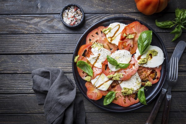 Italian caprese salad with sliced tomatoes, mozzarella cheese, basil, olive oil. Served on black plate over dark wood background. Top view. Rustic style