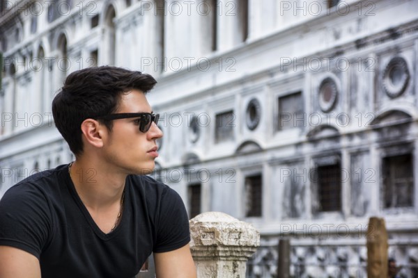 Portrait of Attractive Dark Haired Young Man Leaning Against Railing on Foot Bridge Over Narrow Canal in Venice, Italy