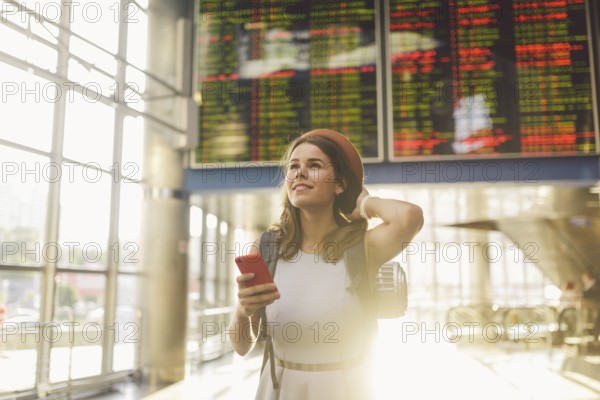 Theme travel and tranosport. Beautiful young caucasian woman in dress and backpack standing inside train station or terminal looking at a schedule holding a red phone, uses communication technology
