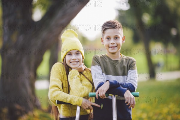Caucasian children twins boy and girl schoolchildren age pose together in sunny weather in autumn park after riding on kick scooters. Happy kids riding kick scooters in public park. Kids sport
