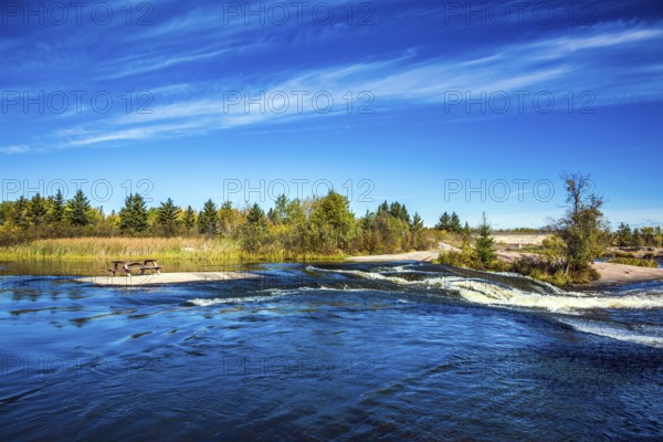 Thin flying cirrus clouds and broad Winnipeg River. Landscape in the Old Pinawa Dam Park. Trend of travel Around the World. On the shore is a wooden bench for tourists