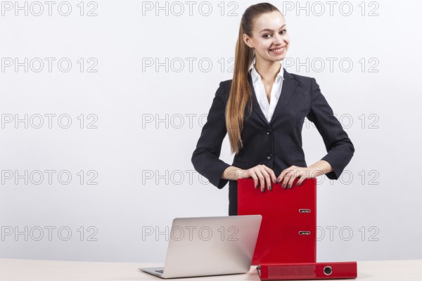 Attractive young caucasian businesswoman with ponytail in black suit stand near the office desk with laptop and hold red paper folders in hands, relies on folders Isolated on grey