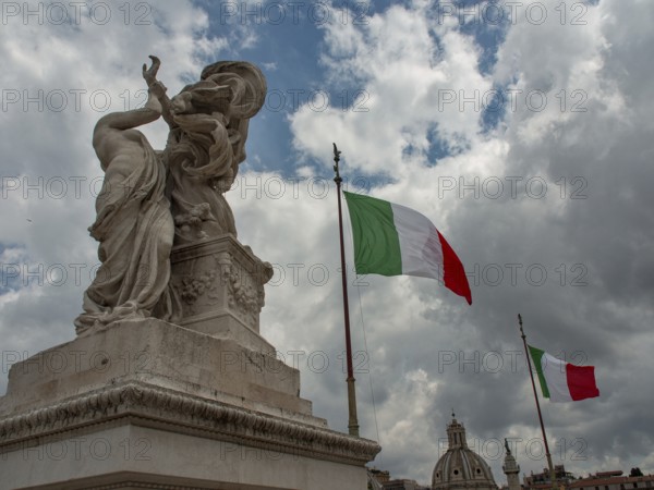 A statue and an Italian flag stand against a dramatic backdrop of clouds with ancient architecture in the background, rome, italy