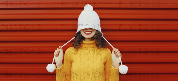 Winter christmas portrait of happy cheerful young woman smiling having fun in yellow sweater, white hat standing on red background