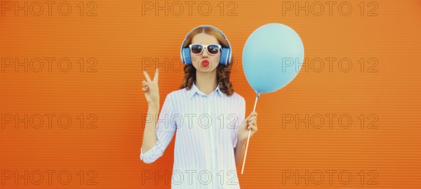 Portrait of happy cheerful young woman in wireless headphones listening to music with balloon on orange background