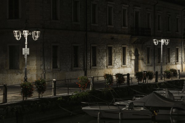 A canal view at night with moored boats, a stone building, and lampposts creating tranquil shadows, night photos taken at long exposure time taken in Peschiera del Garda, Italy Verona