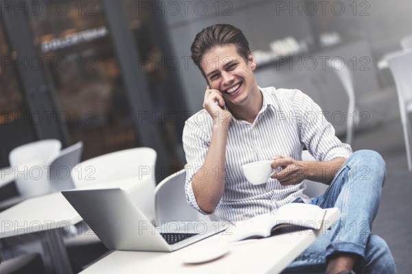 Handsome caucasian young man with phone working on laptop and smiling while enjoying coffee in cafe