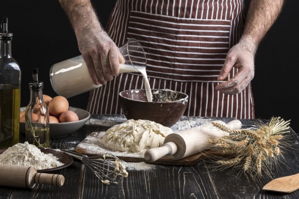 Baker in uniform pouring milk from plastic bottle into bowl. A handful of flour with egg on a rustic kitchen. Ingredients for cooking flour products or dough bread, muffins, pie, pizza dough . Copy space