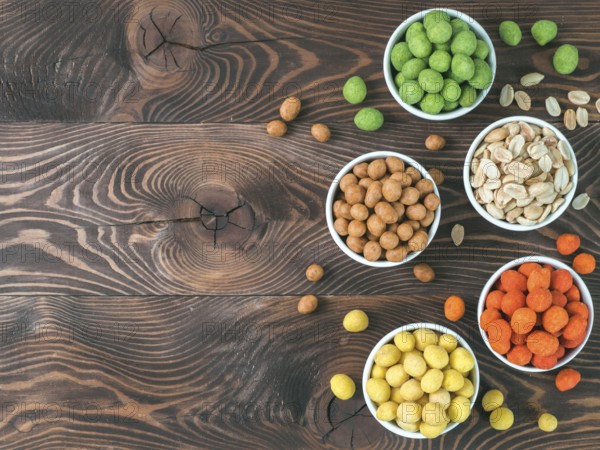 Assortment of different peanuts snack for beer, wine, party bar. Salted peanuts, peanuts in coconut glaze, green vasabi, red spicy chilli, yellow cheese glaze on brown wooden table. Copy space