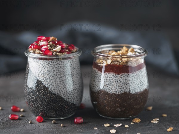 Two chia pudding in glass jars on dark table. Two colors charcoal chia puding with pomegranate, almond, hemp seeds. Chocolate chia pudding with jam and granola. Copy space. Healthy overnight breakfast