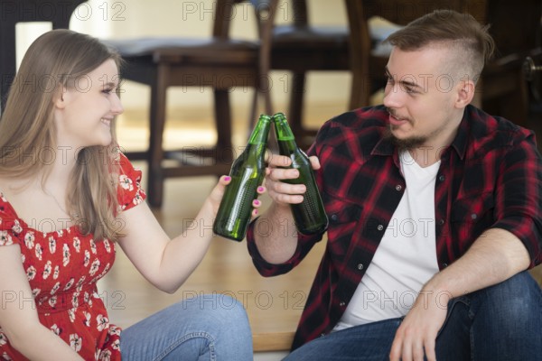 Smiling young people drink beer at a house party. They tap green bottles and look at each other. A wooden table and chairs can be seen in the background