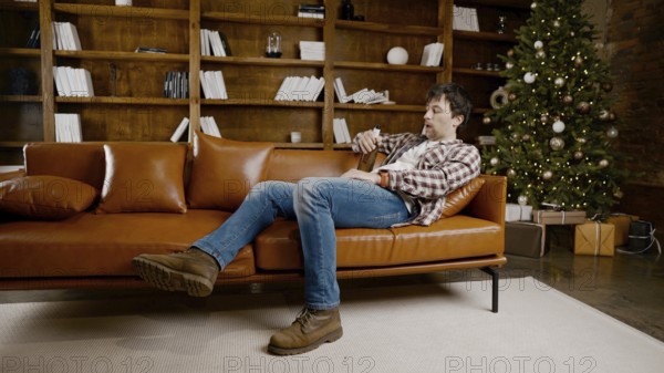 Depressed young man drinking alcohol at home. Unemployed male, drunk and continues drink wine straight bottle on couch near Christmas tree. Addiction, loneliness, nervous breakdown new year holiday