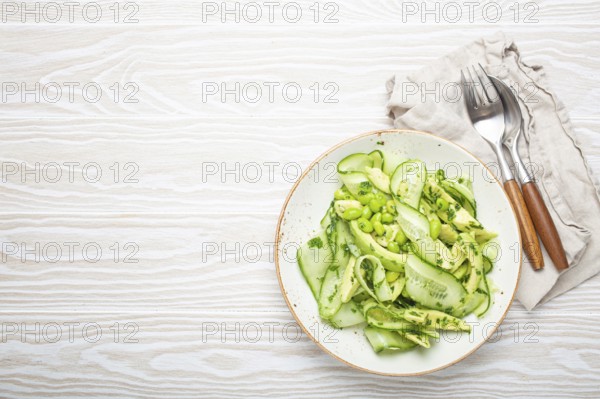 Healthy vegan green avocado salad bowl with sliced cucumbers, edamame beans, olive oil and herbs on ceramic plate top view on white wooden rustic table background. Space for text, food photography, food photography