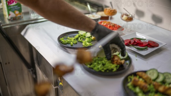 Chef wearing black gloves is garnishing plates with fresh vegetables and sauces, preparing various dishes in a restaurant kitchen specializing in indian cuisine