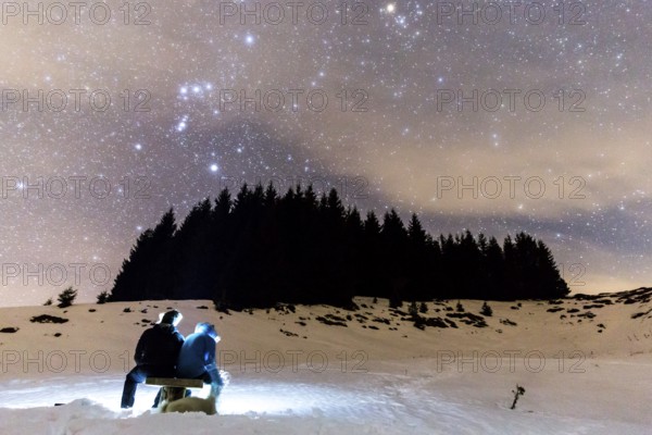 The Milky Way over the winter mountain landscape with pine trees. Two people sitting in the foreground watching the falling stars