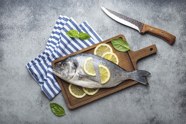 Raw fish dorado in casserole dish with ingredients lemon, fresh basil, bottle of olive oil on wooden cutting board with knife on rustic stone background top view, cooking healthy fish dorado concept, food photography, food photography