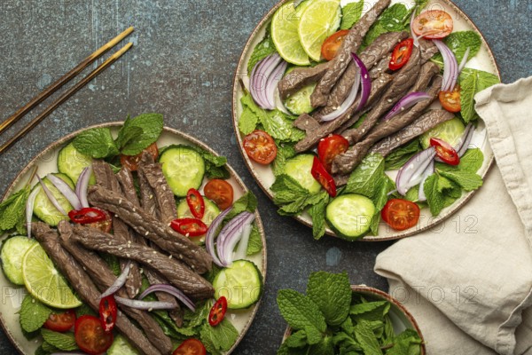 Two plates with traditional Thai beef salad with vegetables and mint top view served on rustic concrete background, healthy exotic asian meal