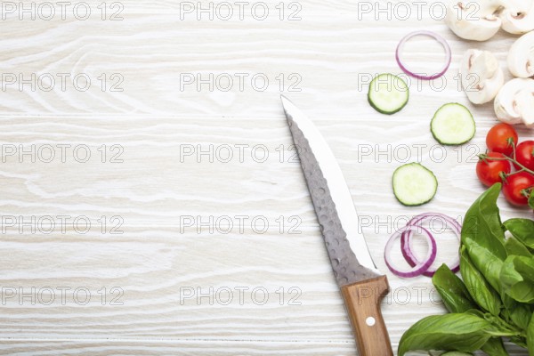 Food cooking background: fresh ingredients with kitchen knife, basil, cherry tomatoes, mushrooms, cut cucumber and onion on white wooden background top view. Meal preparing template, space for text, food photography