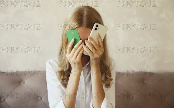 Desperate upset young woman holding mobile phone and bank card touching her head and covering face, internet fraud, problems with online payments or blocked credit card, sitting on the couch at home