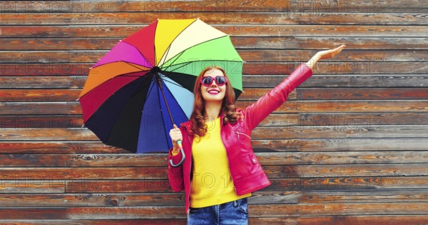 Autumn portrait of happy smiling young woman holding colorful umbrella wearing red jacket, heart shaped sunglasses on city street on wooden wall background