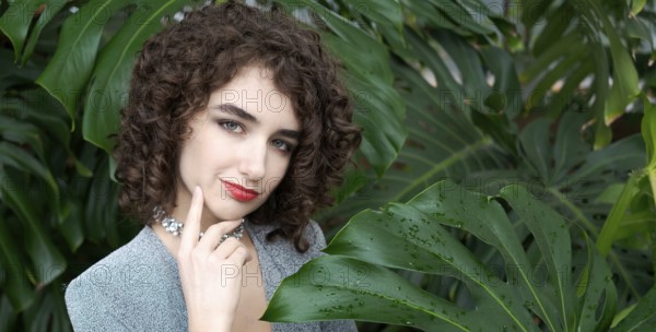 Woman with curly hair is standing in front of a green plant. She is wearing a blue dress and a necklace