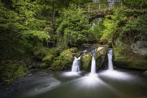 Three small waterfalls under a wooden bridge, surrounded by green vegetation, Mullerthal Region, Luxembourg Switzerland, Grand Duchy of Luxembourg