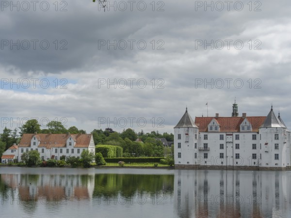 A historic castle with white facades and red roofs is reflected in a calm body of water under a cloudy sky, Glücksburg, Schleswig-Holstein, Germany