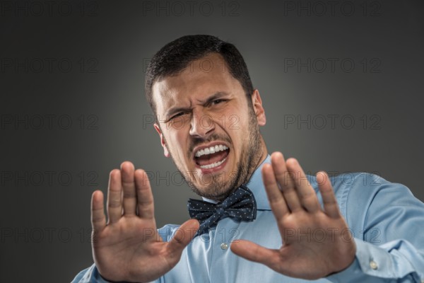 Portrait of angry upset young man in blue shirt and butterfly tie with hands up yelling isolated on gray background. Negative human emotion, hands show Stop, facial expression. Closeup