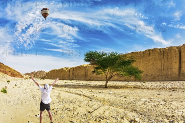 The huge multi-colored balloon flies over desert near the seaside resort of Eilat. The tourist in white t-shirt is delighted with desert