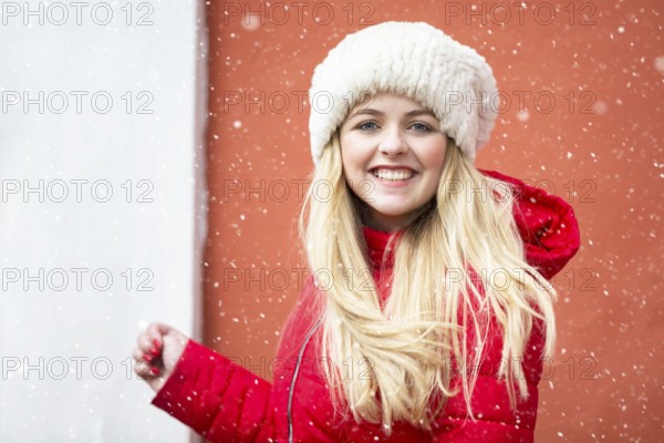 Portrait of a beautiful blonde girl in a warm hat and red jacket and falling snow