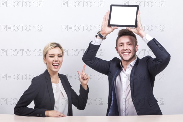 Attractive young caucasian businesswoman with ponytail in black suit and businessman in black suit sit at the office desk and hold empty tablets, cheerful Isolated on grey
