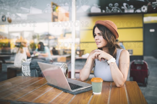 Beautiful young caucasian woman sitting on a terrace in a cafe in summer at a wooden table in a hat and a plate uses technology, working behind a laptop booking hotels trip in the summer