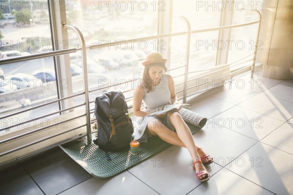 Waiting, delayed transport in the terminal of the airport or train station. Young caucasian woman in dress and hat sits on tourist rug with backpack near window to room and looks into tourist map