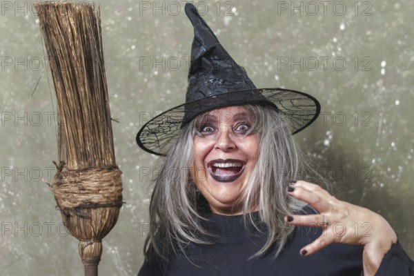 Old witch with white hair and a black hat with an ancient broom in her hands and a terrifying gesture