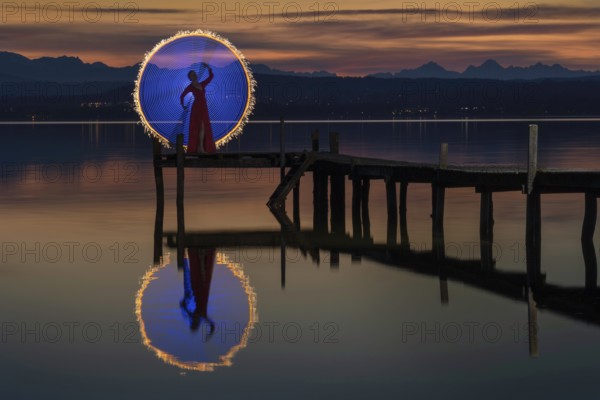 Woman in a red dress on a jetty at sunset on a lake, standing in front of a circle of light created by light painting, Lake Ammer, district of Starnberg, Bavaria, Germany