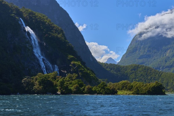 Majestic waterfall flows from forested mountain into the water under a clear blue sky, summer, Milford Sound, Te Anau, Fiordland National Park, South Island, New Zealand